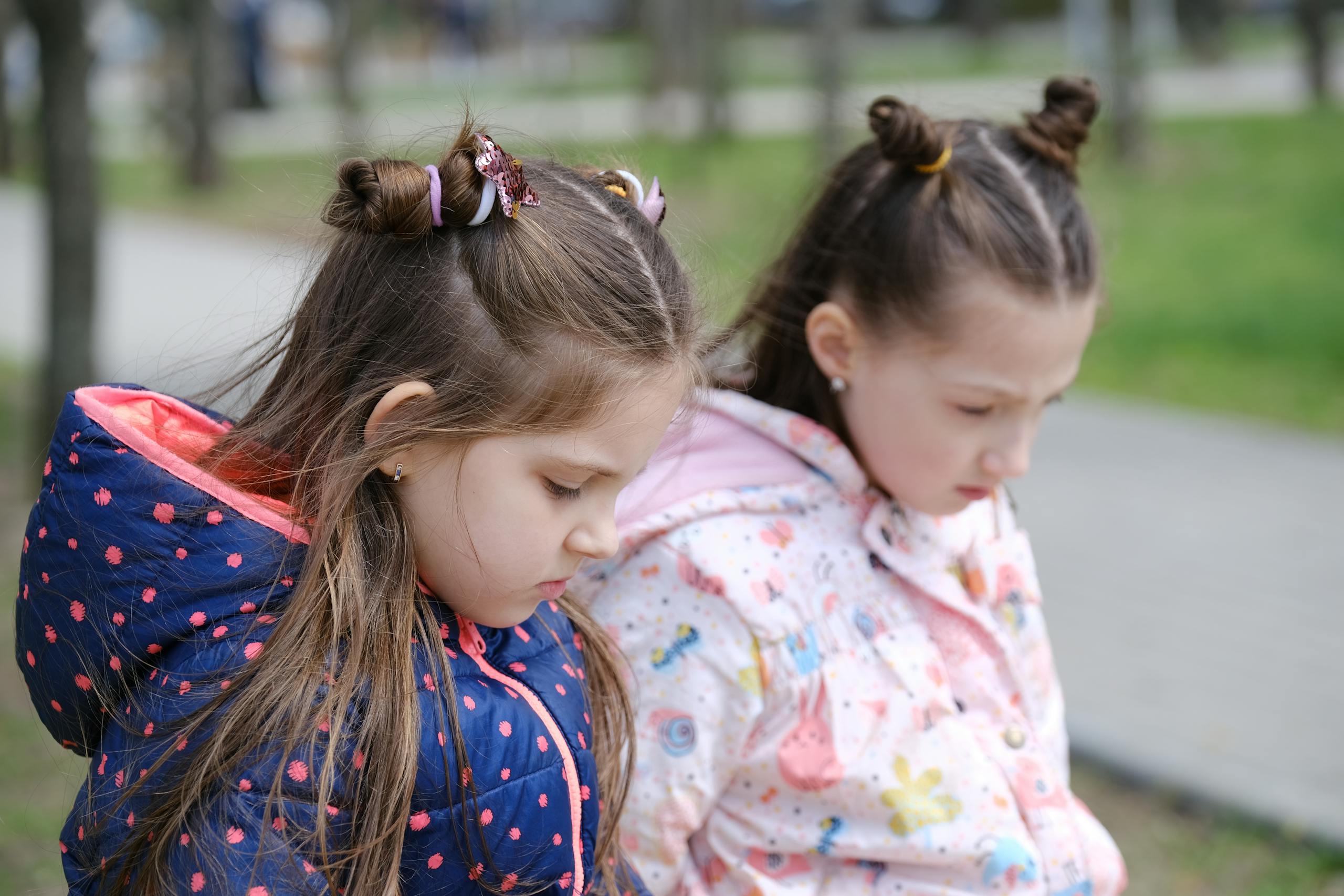 Two young girls in winter jackets, contemplative in a park setting.